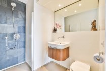 Fregadero en la pared con espejo cerca de inodoro blanco en el cuarto de baño elegante luz con ducha y flores rosas decoradas en el apartamento - foto de stock