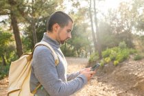 Side view of concentrated young bearded ethnic male traveler in casual clothes and backpack messaging on smartphone standing in lush green forest during trekking in mountainous valley — Stock Photo