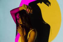 Mujer hispana joven de moda con el brazo levantado mirando hacia otro lado contra la sombra y las luces coloridas del proyector sobre fondo gris - foto de stock