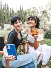 Entzückte multirassische beste Freundinnen mit Cocktailgläsern, die im Sommerpark chillen und ein Selfie mit dem Smartphone machen — Stockfoto