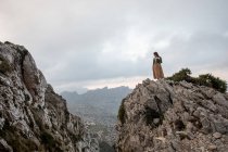 Ganzkörper-Seitenansicht einer verträumten Frau in altmodischer Kleidung, die am Rande einer steinigen Klippe in der Nähe des Meeres bei nebligem Wetter steht — Stockfoto