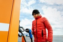From below of thoughtful male in warm outwear and hat standing with charging power to electric vehicle in hand at gas station in sunny day — Stock Photo