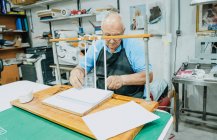 Attentissimo artigiano senior maschio in grembiule e occhiali nastri leganti su tavola di legno prima di lavorare sulla macchina da stampa — Foto stock