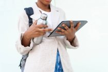 Crop anonyme afro-américaine étudiante avec sac à dos et thermos de boisson chaude surf internet sur tablette sur fond blanc — Photo de stock