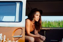 Atractiva chica morena con sombrero dentro de una furgoneta vintage y sentada en el asiento en un día soleado - foto de stock