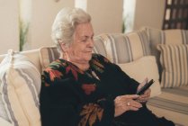 Mujer mayor enfocada con cabello gris descansando en el sofá y leyendo e libro en la tableta en la sala de estar en casa - foto de stock