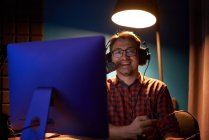 Vue latérale d'un jeune homme concentré en chemise à carreaux et lunettes à l'aide d'un ordinateur et parlant en micro tout en enregistrant un podcast en studio sombre — Photo de stock