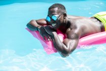 Безстрашний афроамериканець у сонячних окулярах відпочиває на рожевому надувному матраці в басейні під час купання сонця в сонячний літній день. — стокове фото
