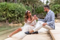 Full length of positive married couple in wedding outfit and sunglasses sitting on stone stair near lake and green palms and plants while looking at each other and giving flower — Stock Photo