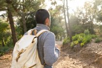 Back view of unrecognizable bearded male hiker in casual clothes and backpack standing in green forest on sunny day during trekking — Stock Photo