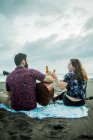 Vista posteriore di una coppia positiva di musicisti seduti con chitarre e bottiglie di birra mentre sono seduti sulla spiaggia sabbiosa vicino all'oceano durante il giorno — Foto stock