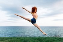 Ganzer Körper der barfüßigen Frau beim Split, während sie mit erhobenen Armen an der Küste des plätschernden Meeres hoch springt — Stockfoto