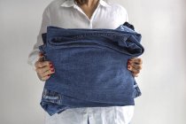 Crop anonimo femminile in camicia bianca con pila di jeans blu in mano — Foto stock
