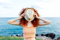 Unrecognizable female with long ginger curls covering face with hat on coast of blue sea — Stock Photo