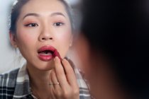 Cosecha joven atractiva hembra asiática en ropa casual aplicando lápiz labial brillante mientras mira el espejo - foto de stock