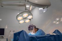 Professionista senior chirurgo maschile in maschera e uniforme facendo operazione sotto lampada in sala operatoria — Foto stock