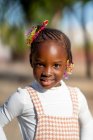 Glückliches afroamerikanisches Mädchen mit Frisur in stilvollem Outfit steht und schaut in die Kamera vor verschwommenem Hintergrund auf der Straße an sonnigen Tagen — Stockfoto