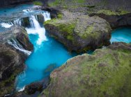Сверху стремительный каскад падает в голубом озере в окружении грубых скалистых образований, покрытых зеленым мхом в природе Исландии — стоковое фото