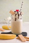 Стеклянная банка сладкого бананового сплит молочного коктейля с шоколадом со взбитыми сливками и вишней на разделочной доске — стоковое фото