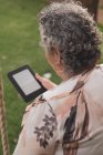 Visão traseira da senhora idosa vestindo blusa sentada no parque e lendo livro eletrônico — Fotografia de Stock