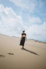 Vista trasera de cuerpo completo de una hembra descalza irreconocible en vestido paseando sobre una duna arenosa en el desierto - foto de stock