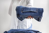 Обрізати анонімну жінку в білій сорочці зі стопкою синіх джинсів в руках — стокове фото