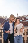 Улыбающийся бородатый мужчина с бутылкой пива обнимает позитивную девушку прокручивая мобильный телефон на балконе в солнечный день — стоковое фото