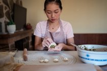 Mulher em roupas casuais e avental recheando bolinhos com carne enquanto prepara jiaozi chinês tradicional na cozinha — Fotografia de Stock