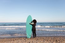 Visão traseira de surfista feminina irreconhecível em roupa de mergulho com prancha de surf de pé olhando para longe na praia lavada pelo mar ondulando — Fotografia de Stock