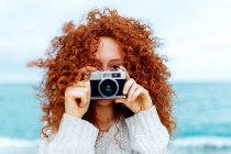 Ginger haired female in knitted sweater looking at camera while taking picture on retro photo camera on coast of sea — Stock Photo