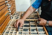 De arriba de la cosecha concentrado senior macho artesano en delantal y anteojos elegir impresión prensa letras de madera caja durante el trabajo en tradicional atelier - foto de stock