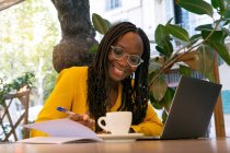 Lächelnde afroamerikanische Freelancerin macht sich Notizen im Notizblock, während sie mit Netbook und Tasse Drink am Tisch sitzt, während sie in der Cafeteria fernarbeitet — Stockfoto