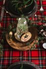 Cenário de mesa de Natal com coroa de flores e ornamentos decorativos de madeira e toalha de mesa quadriculada vermelha com luzes amarelas no fundo — Fotografia de Stock