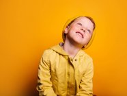 Niño feliz en chaqueta de impermeable amarillo vivo y gorro sombrero riendo y mirando hacia otro lado contra el fondo amarillo en el estudio - foto de stock