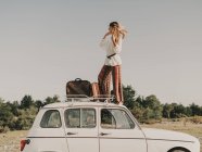 Вид всего тела сбоку стильной женщины-хиппи в одежде, стоящей на белом старом автомобиле с багажником во время поездки на природу — стоковое фото