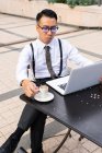 Jeune entrepreneur asiatique poignant avec tasse de boisson chaude et netbook regardant l'écran dans la cafétéria urbaine table en plein jour — Photo de stock