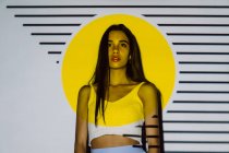 Созерцательная молодая латиноамериканка с макияжем и длинными волосами, смотрящая в желтый проектор света с полосками — стоковое фото