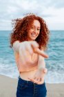 Mulher agradável com penteado encaracolado de gengibre longo que alcança a mão à câmera ao estar contra o mar ondulando — Fotografia de Stock