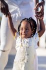 Happy African American little girl in light dress holding hands of unrecognizable father while walking on street in sunny day — Stock Photo