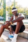 Happy African American male basketball player sitting on sports ground and browsing smartphone on sunny day in summer — Stock Photo