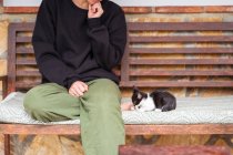 Обрізана невпізнавана молода етнічна жінка, що взаємодіє з чарівною кошеням, сидячи з схрещеними ногами на лавці — стокове фото