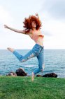 Corpo inteiro de mulher enérgica feliz com cabelo encaracolado de gengibre voador pulando acima do chão gramado na costa do mar ondulante azul olhando para longe — Fotografia de Stock