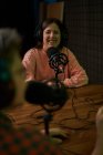 Молодая радиоведущая в повседневной одежде и наушниках сидит за столом с микрофоном и общается с анонимным коллегой во время записи подкаста в студии — стоковое фото