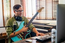 Männlicher Gitarrist mit Brille und Kopfhörer, der Instrument hält, während er Computer im modernen Studio benutzt — Stockfoto