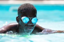 Спокойный афроамериканец в стильных солнцезащитных очках с отражением водного плавания в бассейне на размытом фоне в солнечный летний день — стоковое фото