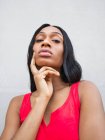 Mulher afro-americana confiante com cabelo preto tocando na bochecha e olhando para a câmera enquanto estava em pé no fundo branco na rua — Fotografia de Stock