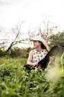 Спокійна жінка-музикантка в повсякденному одязі сидить на зеленій траві і відкриває чорний випадок акустичного укулеле в денне світло — стокове фото