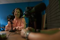 Feliz presentadora de radio femenina en ropa casual y auriculares sonriendo mientras graba podcast en estudio moderno con colega irreconocible de cosecha - foto de stock