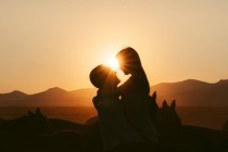 Side view of silhouettes of loving couple hugging each other while spending time together on pasture near horses against mountains in sunset — Stock Photo