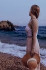 Donna deliziata con i capelli lunghi in abito alla moda in piedi sulla riva del mare in estate sera mentre tiene il cappello con una mano — Foto stock